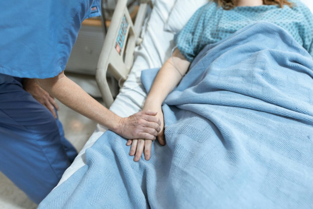 A nurse in scrubs offers comfort by holding a patient's hand in a hospital bed.